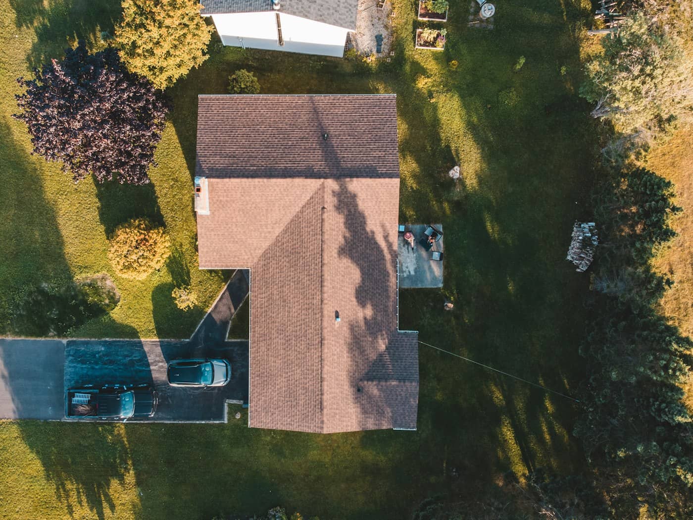 Drone top-down of a home's shingle roof at golden hour — ridge, hips, and chimney casting long evening shadows across warm brown courses