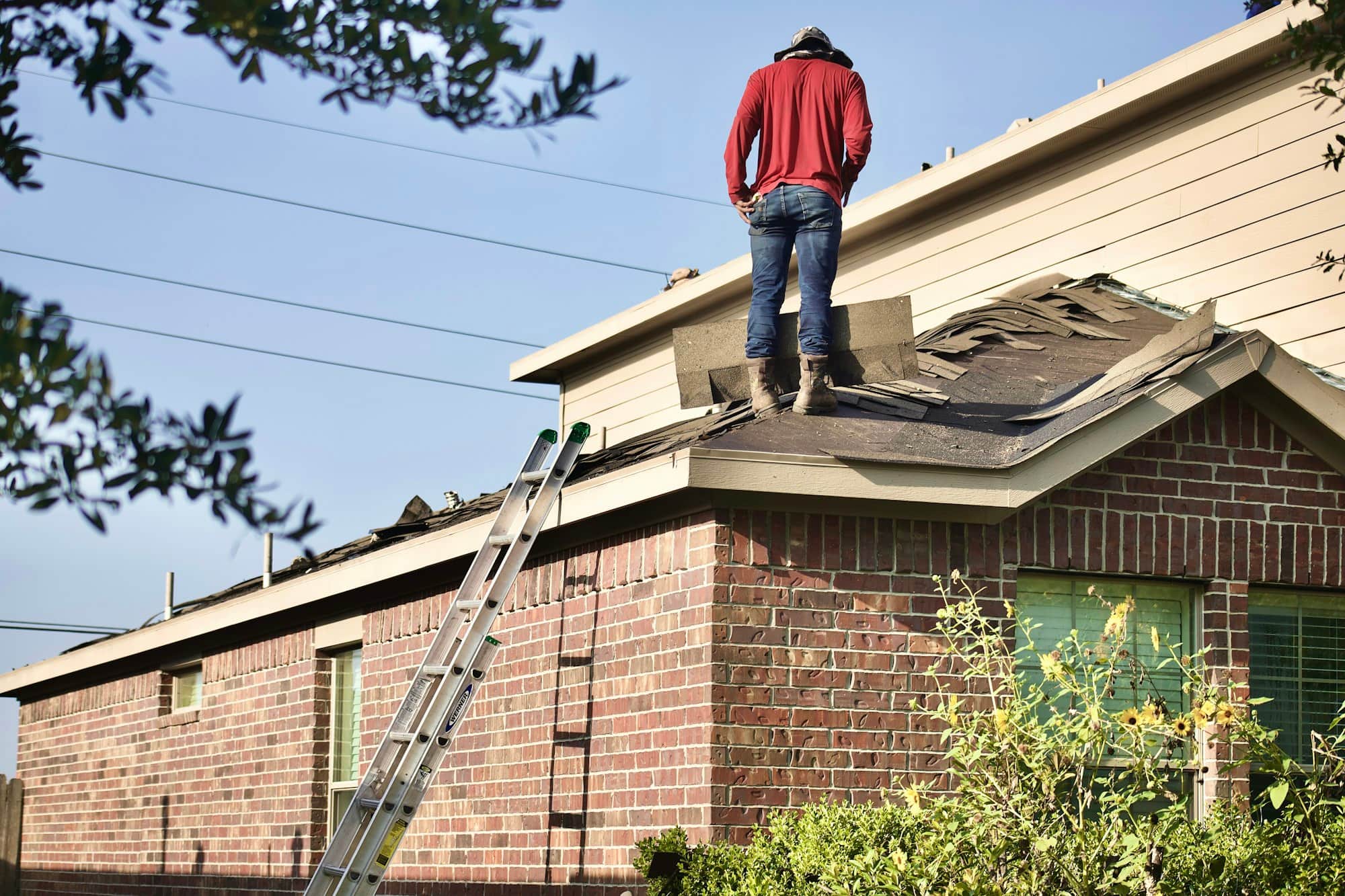 Roofer standing on a brick Fort Worth home mid tear-off, shingles stripped to the decking under a clear Texas sky