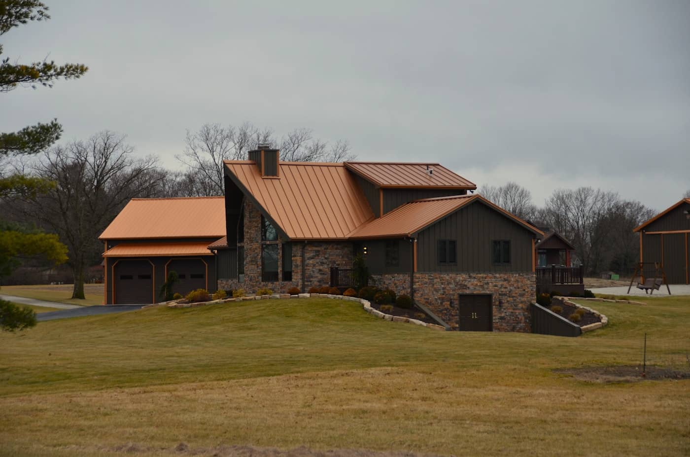 Copper-toned standing-seam metal roof over a stone-and-timber home — clean vertical seams, concealed clips, no exposed fasteners